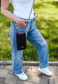 Black crossbody bag with a pink zipper, featuring a chain strap. Worn with light blue distressed jeans and white sneakers. Green grass background.