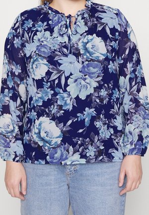 Femme portant un chemisier bleu à fleurs à manches longues avec un col froncé et un jean bleu clair, debout sur un fond uni.