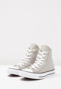Hoge sneakers met een lichtgrijze mesh bovenkant, een witte rubberen zool en zwarte details, met witte veters en een gestructureerd oppervlak.