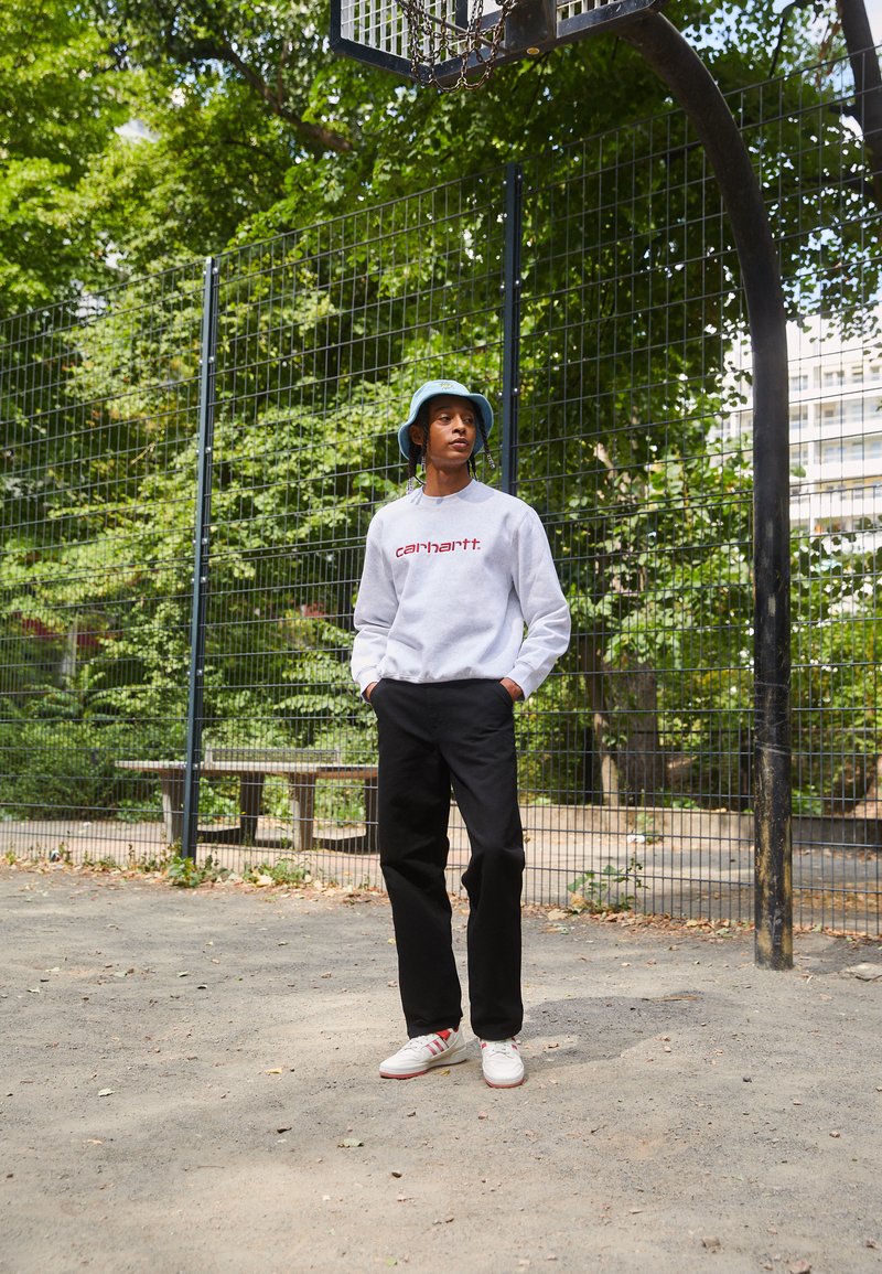 Grauer Sweatshirt mit rotem "Carhartt"-Logo, schwarze Hose und weiße Sneaker. Die Person trägt einen blauen Bucket Hat und steht in der Nähe eines Basketballkorbs.