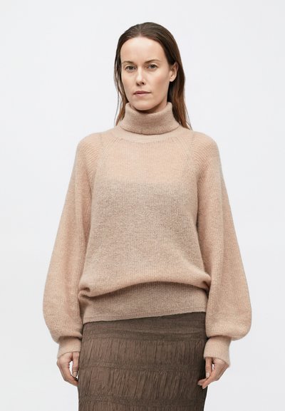 Világos rózsaszín kötött pulóver magas nyakkal és széles ujjakkal, puha tapintású textúrával. Barna, redős szoknyával párosítva.