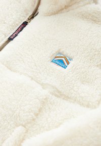 Záber zblízka na krémovú fleecovú bundu s nášivkou loga značky Superdry a sťahovaním zipsu.