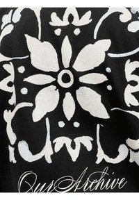 Tissu noir avec un grand motif floral blanc comprenant six pétales et des éléments décoratifs symétriques, avec « Ours Archive » en écriture cursive en dessous.