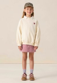 Sweatshirt crème avec un patch en forme de cœur, jupe en velours côtelé rose, chaussettes à motifs et chaussures marron. La casquette a un motif léopard.