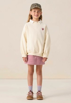 Crème sweatshirt met een hart patch, roze corduroy rok, gemusterde sokken en bruine schoenen. Pet heeft een luipaardprint.