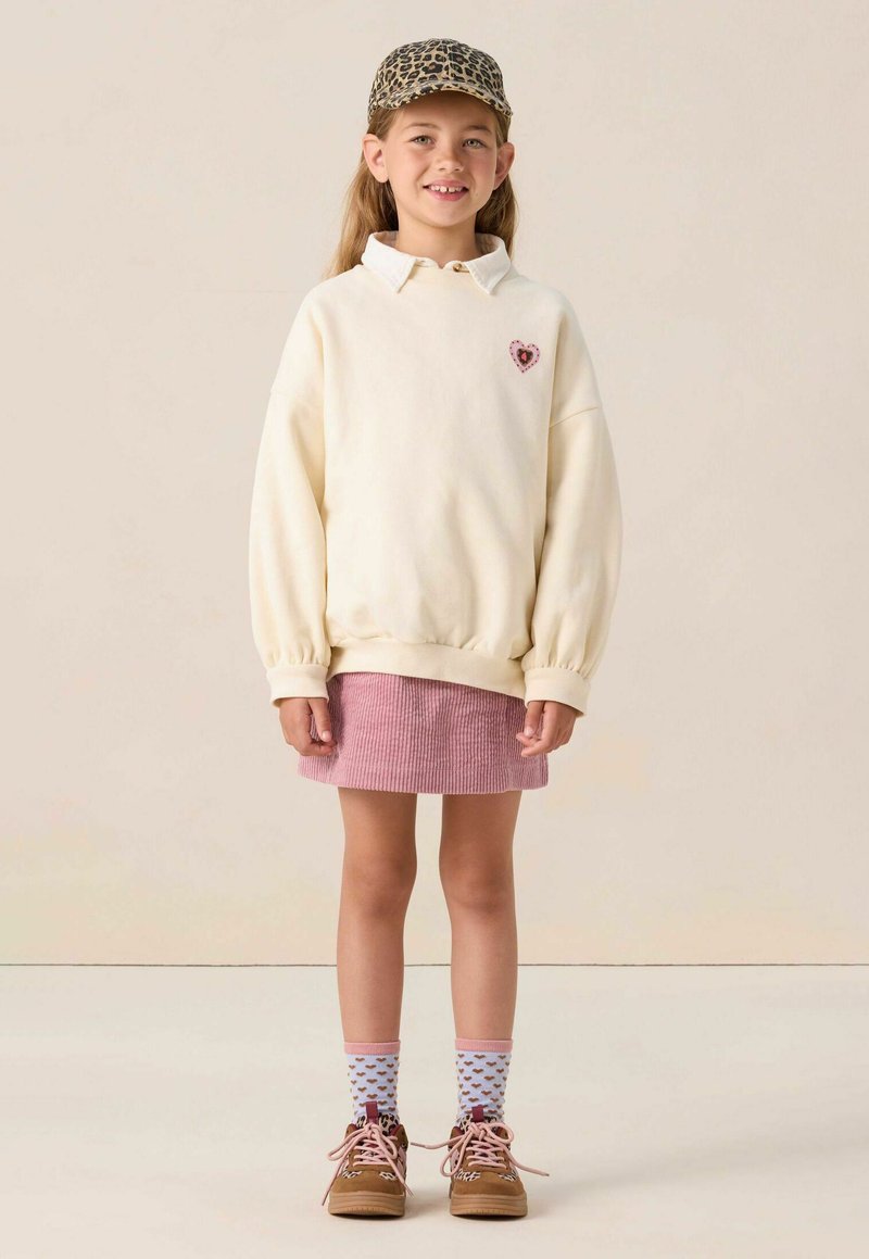 Sweatshirt crème avec un patch en forme de cœur, jupe en velours côtelé rose, chaussettes à motifs et chaussures marron. La casquette a un motif léopard.