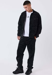 Giovane che guarda in basso, indossando una giacca e pantaloni di velluto a coste neri, una maglietta bianca con logo e scarpe sportive nere su sfondo neutro.