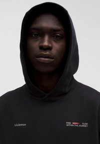 Person in einem schwarzen Kapuzenpullover mit hochgezogener Kapuze, auf der Brust ist der Text "lululemon" und "Finde den Fluss in der Reise" auf einem einfarbigen Hintergrund.