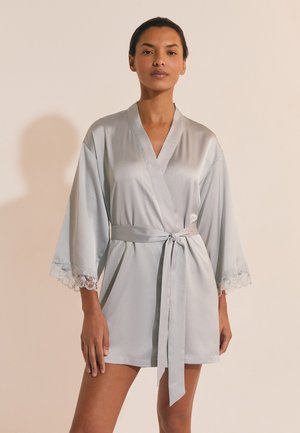 Donna che indossa un accappatoio di raso grigio chiaro con maniche rifinite in pizzo annodato in vita, in piedi contro uno sfondo beige.