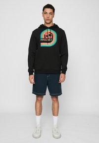 Fekete kapucnis felső elülső grafikával narancssárga, kékkel és sárgával, pálmafák és "ROLLER SKATER CLUB VENICE BEACH" felirattal. Sötét rövidnadrággal párosítva.