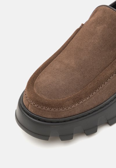 Braunes Wildleder Slipper mit einer dicken schwarzen Gummisohle, ausgestattet mit einer abgerundeten Zehenpartie und erhöhten Nähten entlang des Kragens.
