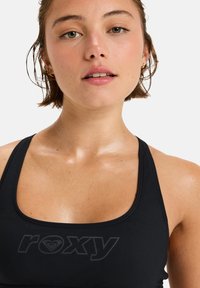 Black sports bra with a rounded neckline, featuring a subtle Roxy logo in dark print. Material appears smooth and fitted.