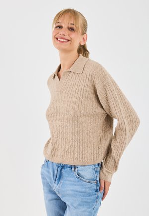 Beige Pullover aus Kabelstrick mit Kragen und langen Ärmeln, der ein strukturiertes Muster aufweist. Getragen mit hellblauen Jeans.