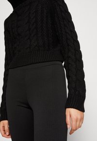 Schwarzer Strickpullover mit Rippensaum und langen Ärmeln, kombiniert mit schwarzen Rippleggings. Glatte Textur mit einer taillierten Silhouette.