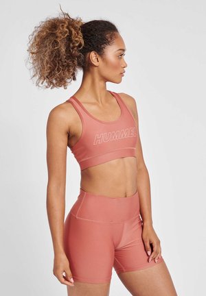 Fit kvinde med krøllet hestehale iført matchende koral sportstop og shorts, der kigger til højre mod en ensfarvet baggrund.