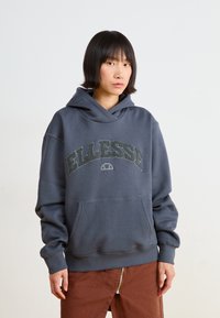 Felpa grigia con cappuccio e grande logo "ELLESSE" in rilievo. Presenta una tasca frontale e polsini a costine, realizzata in un tessuto morbido.