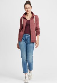 Sweat à capuche zippé bordeaux, avec des cordons de serrage crème, associé à un jean bleu clair et des baskets beiges. Tissu lisse, coupe décontractée, poignets côtelés.