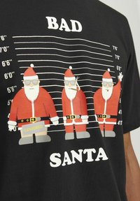 Sort T-shirt med tre tegnede julemander i røde dragter, solbriller og et "CENSURERET" skilt, anordnet i en højdemålsopstilling.