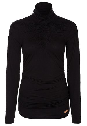 Mujer que lleva una blusa negra de manga larga, pantalones ajustados beige, tacones negros de punta y un bolso negro con cadena al hombro.