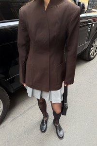 Tmavohnedý blazer so prispôsobeným strihom, s vysokým golierom a dvoma prednými vreckami. V páre s plisovanou šedou sukňou a čiernymi mokasínami.