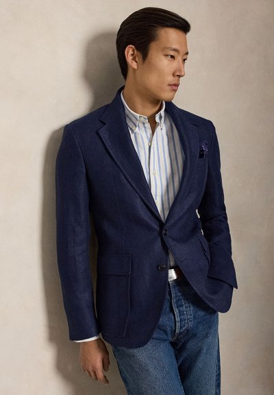 Mann trägt einen marineblauen Blazer, ein gestreiftes Hemd und Jeans, steht mit einer Hand in der Tasche und schaut seitlich vor einer einfachen Wand.
