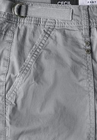 Pantaloni grigi chiari con una texture di tessuto liscia, tasche frontali, passanti per cintura e bottoni metallici. Etichetta del marchio visibile in alto.