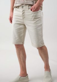 Shorts en denim beige avec des revers roulés, poches avant et taille à bouton. Texture lisse avec un léger délavage et des détails de couture subtils.