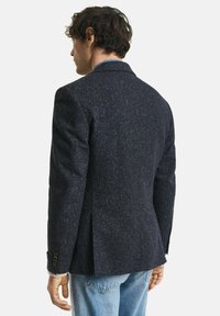 Uomo che indossa un blazer scuro a pois e jeans azzurri chiari, rivolto di spalle su uno sfondo bianco semplice.