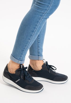 Person wearing rolled-up blue skinny jeans and black Gore-Tex sneakers with white soles, standing on a white surface.