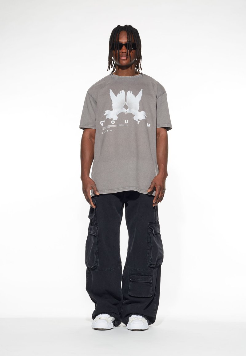 Grå oversize t-shirt med en vit fågelgrafik och texten "LOST YOUTH", kombinerad med lösa svarta cargo-byxor och vita sneakers.