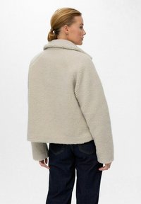 Veste en fausse fourrure beige avec un large col, à design court et surface texturée, associée à un jean bleu foncé, vue de dos.