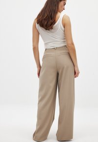Bež hlače širokih nogavic z gladko teksturo, s prilegajočim pasom, zankami za pas in dvema zadnjima žepoma, v kombinaciji z belo majico brez rokavov.