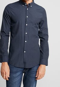 Man med mörkblå skjorta med knapp och ett litet geometriskt mönster, samt blå jeans, stående framför en ljus och enfärgad bakgrund.