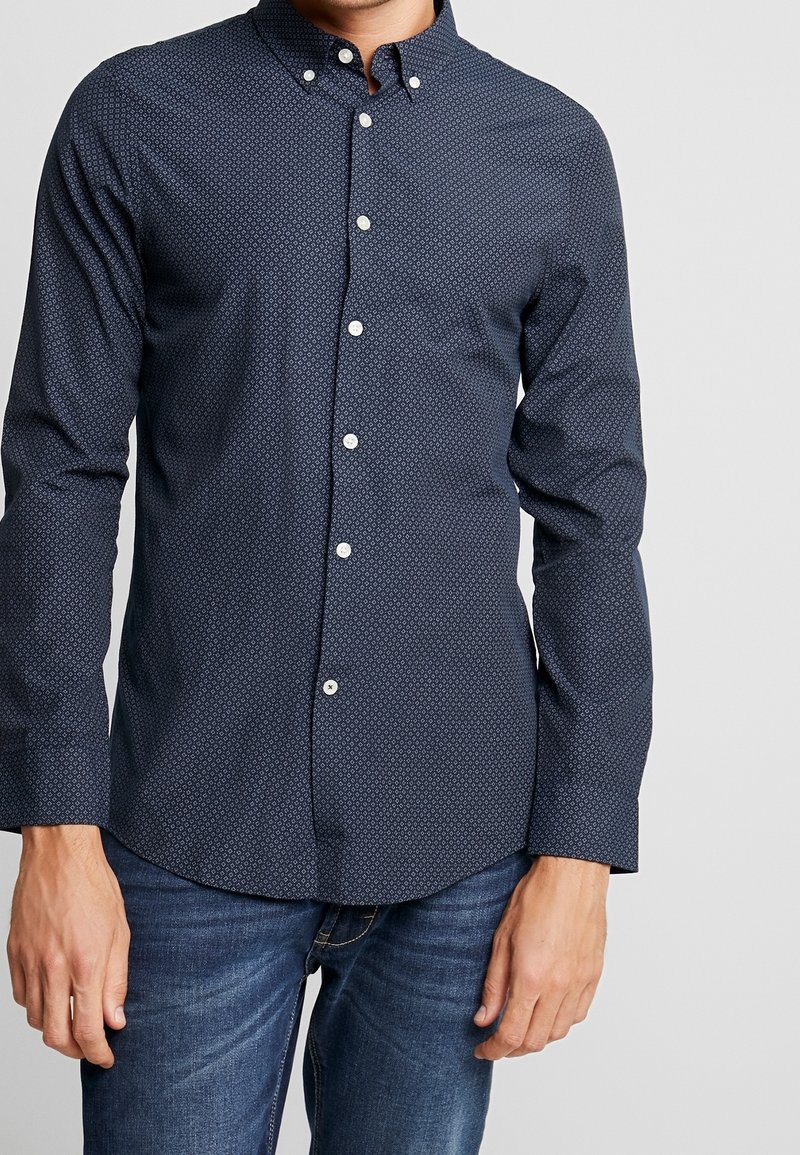 Man med mörkblå skjorta med knapp och ett litet geometriskt mönster, samt blå jeans, stående framför en ljus och enfärgad bakgrund.