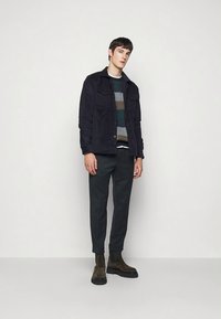 Námořnicky modrá semišová bunda přes víceméně barevný proužkovaný svetr, v kombinaci s tmavými kalhotami a hnědými kotníkovými botami. Minimalistické pozadí.