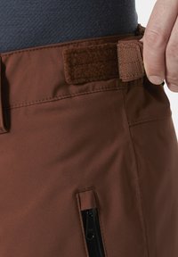 Bruine, zachte stoffen broek met een close-up van de tailleband met een klittenbandsluiting, voorzien van een zwarte ritssluiting aan de zijkant.