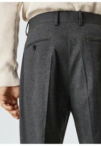Gros plan sur une personne portant un pantalon droit gris foncé avec des poches arrière boutonnées et un pull en tricot beige.