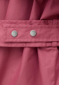 Close-up van een opgevouwen stoffige roze stof met een gestikte riemlus en riem, bevestigd met twee ronde zilverkleurige drukkers.