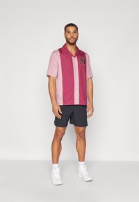 Polo majica s kratkimi rokavi v bordo in svetlo roza barvi z gumbom spredaj in NY logotipom, parjena z črnimi kratkimi hlačami in belimi športnimi copati.
