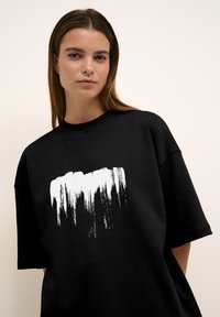 Musta oversized t-paita, joka on valmistettu puuvillasta. Paidassa on valkoinen siveltimen jälki -kuvio rinnassa. Lyhyet hihat ja pyöreä kaula-auko.