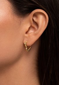 Orecchino a cerchio in oro con design intrecciato, caratterizzato da piccoli accenti sferici e un'aggiunta di cristallo. Indossato all'orecchio di un modello.