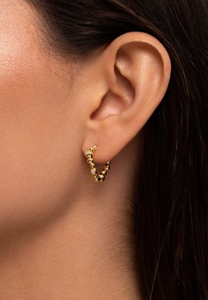 Gouden ringoorbel met een gedraaide design, voorzien van kleine sferische accenten en een kristallen versiering. Draagbaar op het oor van een model.