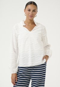Blusa bianca a maniche lunghe con una texture ondulata e bordi strutturati. Presenta un collo a V e polsini arr gathered. Abbinata a pantaloni a righe blu navy.