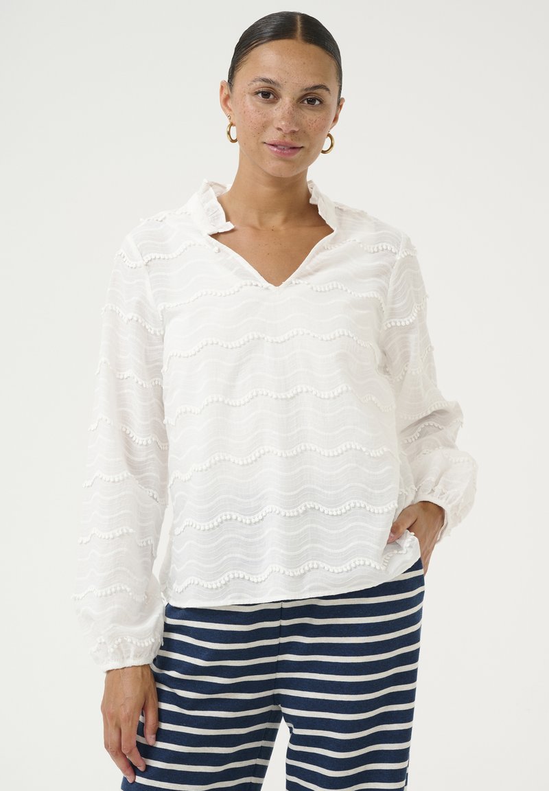 Blusa bianca a maniche lunghe con una texture ondulata e bordi strutturati. Presenta un collo a V e polsini arr gathered. Abbinata a pantaloni a righe blu navy.