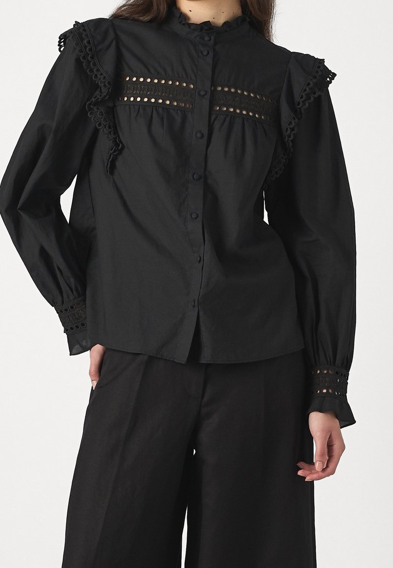 Frau trägt eine schwarze, geknöpfte Bluse mit Spitzenbesatz und Ösen-Details an Brust, Schultern und Manschetten, kombiniert mit schwarzen, weit geschnittenen Hosen.