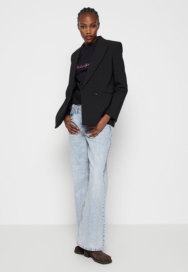 Schwarze doppelt geknöpfte Blazer über einem schwarzen T-Shirt mit pinkem Text, kombiniert mit hellblauen Schlagjeans und braunen Stiefeletten mit Absatz.
