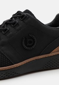 Svart sneaker med en strukturerad läderöverdel, nätackenter, brun gummisula och en framträdande svart logotyp på sidan.