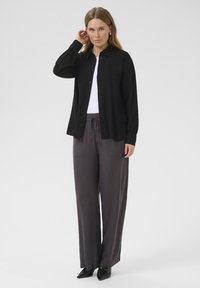Zwarte button-up shirt met lange mouwen en manchet, gedragen over een witte top. Gecombineerd met losse grijze broek met voorzakken en een trekkoord.