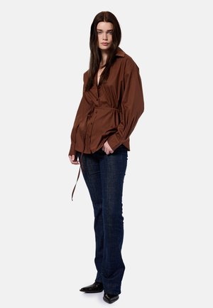Femme aux longs cheveux bruns portant une chemise marron ample boutonnée avec un lien à la taille, un jean bleu foncé et des chaussures noires à bout pointu, debout.
