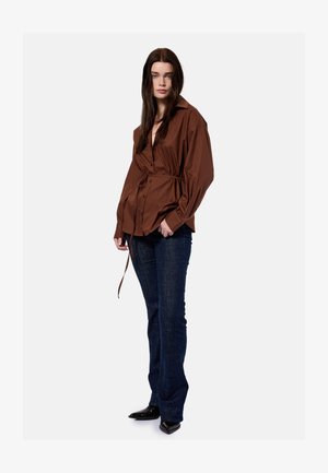 Femme aux longs cheveux bruns portant une chemise marron ample boutonnée avec un lien à la taille, un jean bleu foncé et des chaussures noires à bout pointu, debout.